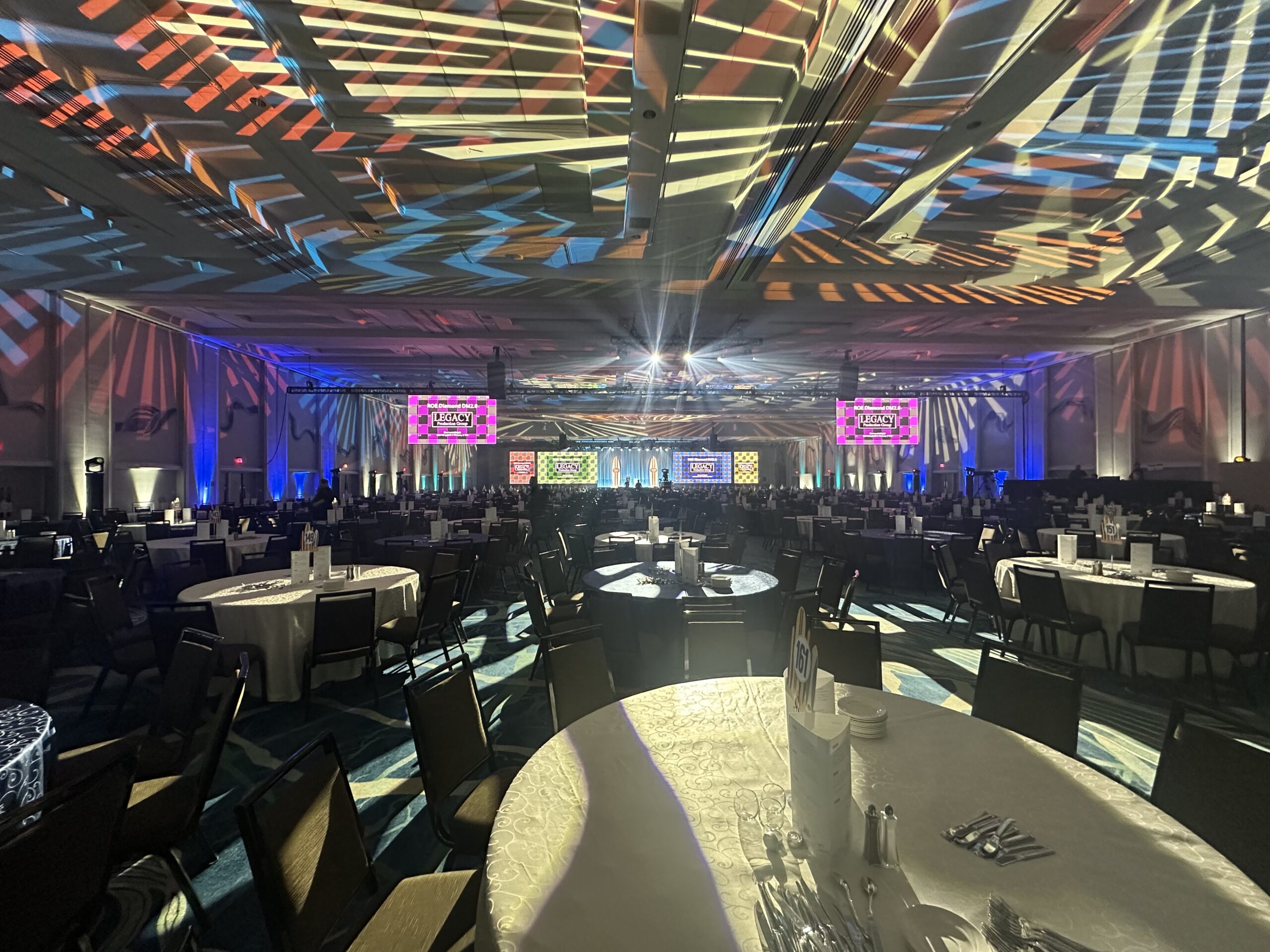 Wide-angle view of a hotel ballroom banquet setup with dozens of round tables dressed in white linens and black chairs under dynamic gobo patterns and blue-and-purple uplighting, all facing a front stage framed by flown truss lighting and multiple colored LED video screens displaying branded graphics, ready for a gala dinner presentation