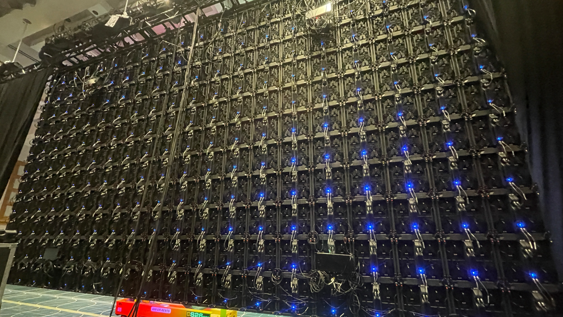Back of Large LED Wall Dark event stage illuminated by a low magenta LED strip at floor level, overhead moving-head lights casting amber and white beams upward, and intricate blue-and-gold gobo patterns projected across the stage floor in a high-impact lighting design