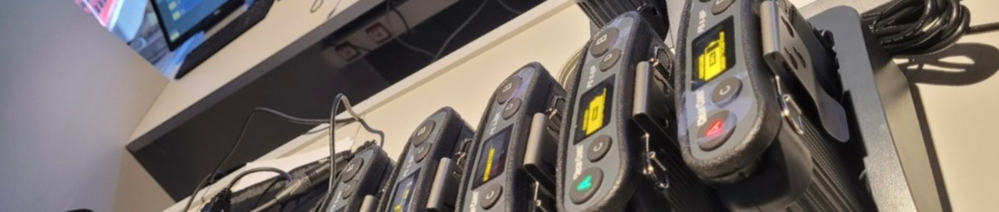Close-up of four wireless bodypack transmitters with belt clips and LCD status displays, neatly lined up on a white conference control desk, illustrating professional wireless audio solutions for live events