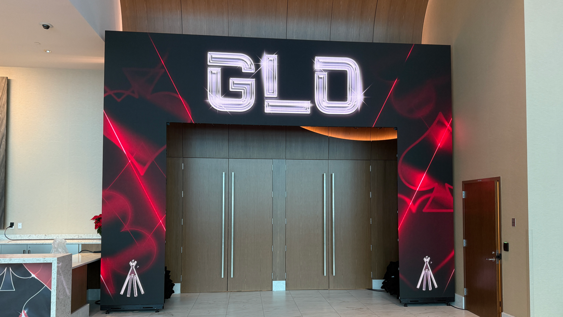 LED Arch over door Modern corporate lounge and meeting room featuring a round conference table with black rolling chairs, two black leather armchairs beside a floor-to-ceiling window, adjustable-height desks with task chairs and red desk lamp, gallery wall prints of AV equipment, and carpeted flooring under recessed ceiling lights