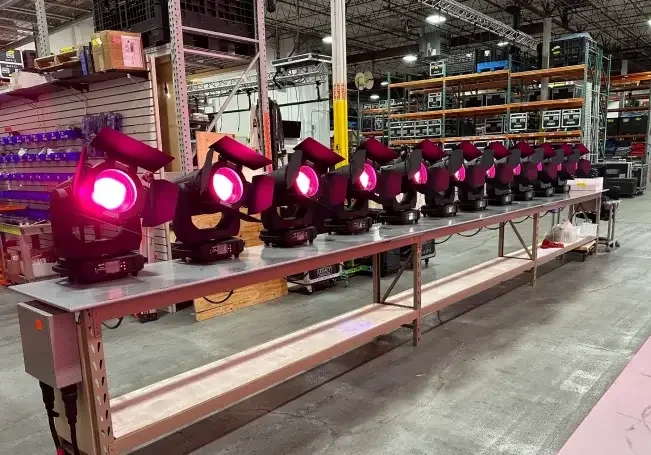 Warehouse staging area with a long metal workbench holding a row of black moving-head stage lights emitting magenta wash; industrial shelving stocked with AV flight cases and truss sections fills the background