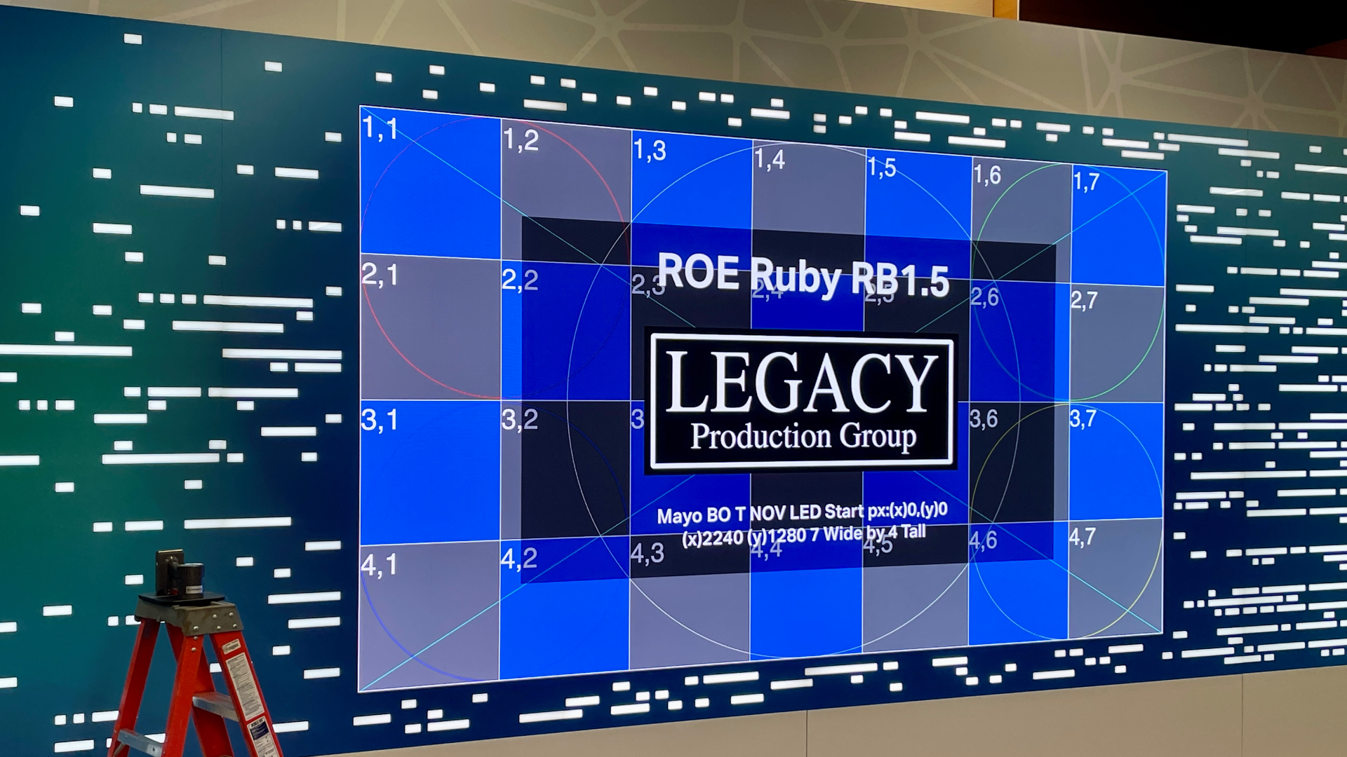 LED Installed Wall Dark event stage illuminated by a low magenta LED strip at floor level, overhead moving-head lights casting amber and white beams upward, and intricate blue-and-gold gobo patterns projected across the stage floor in a high-impact lighting design