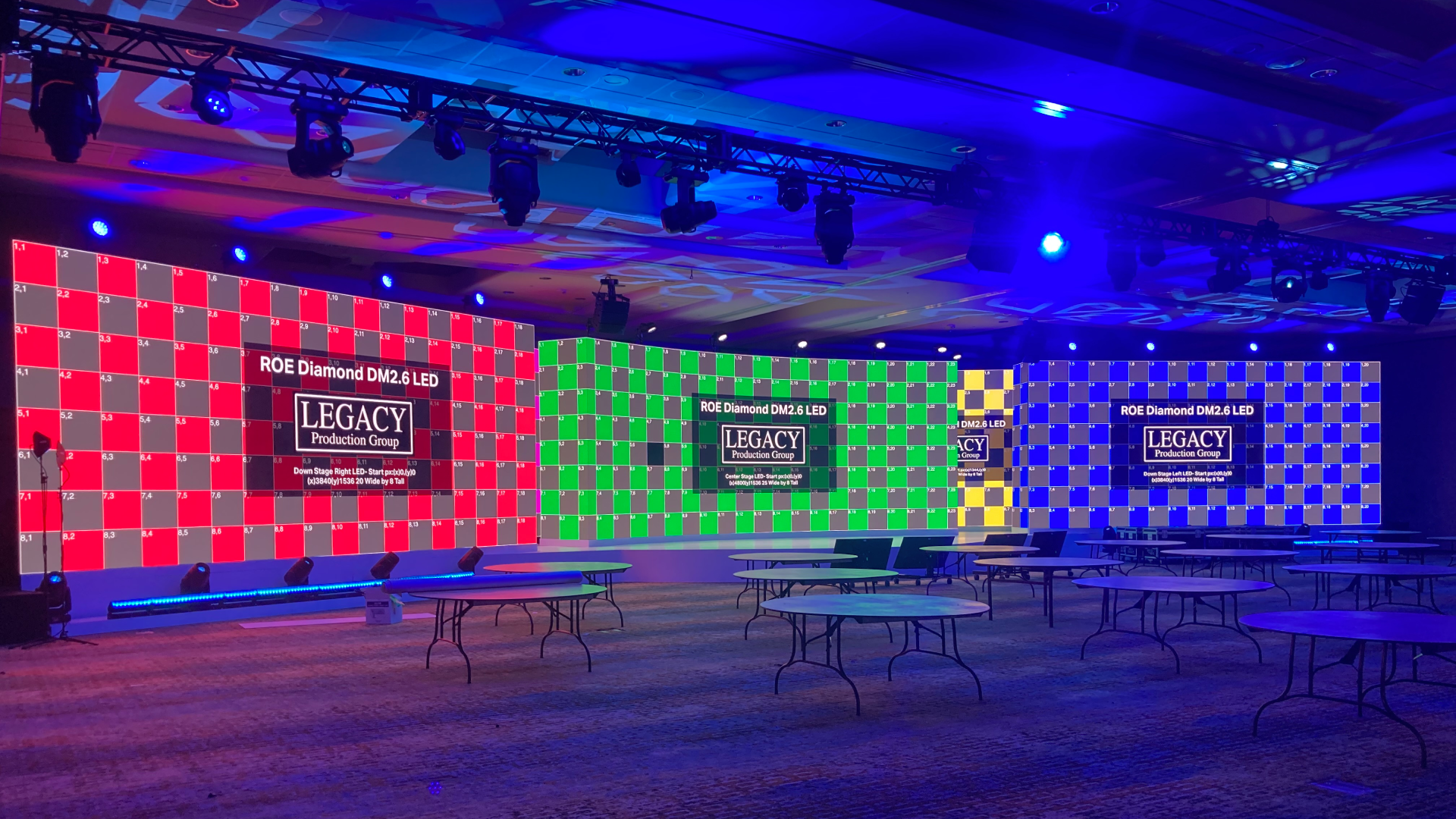 Three very large LED Walls Live production control room with five operators wearing headsets seated at desks loaded with laptops, video switchers, and audio mixers; two large wall-mounted screens display multi-view camera feeds and a bright studio preview in a carpeted office space
