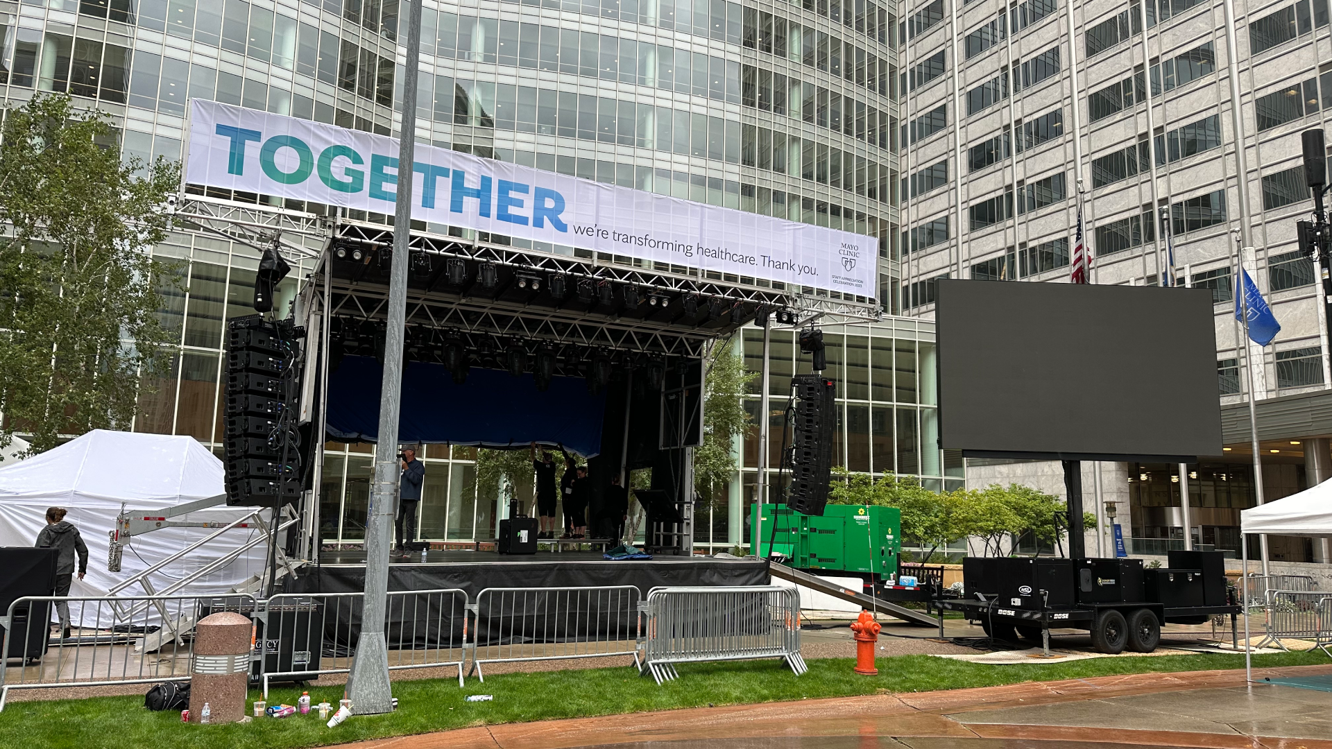 OOutdoor Concert Stage Live production control room with five operators wearing headsets seated at desks loaded with laptops, video switchers, and audio mixers; two large wall-mounted screens display multi-view camera feeds and a bright studio preview in a carpeted office space