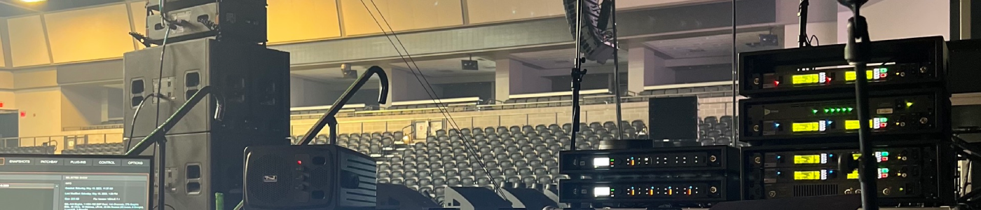 Backstage production station view inside an arena, featuring a video preview monitor at left, a floor monitor speaker beside it, and a rack of wireless microphone receivers with illuminated front panels at right, all overlooking rows of empty seats and overhead lighting trusses ready for live event production