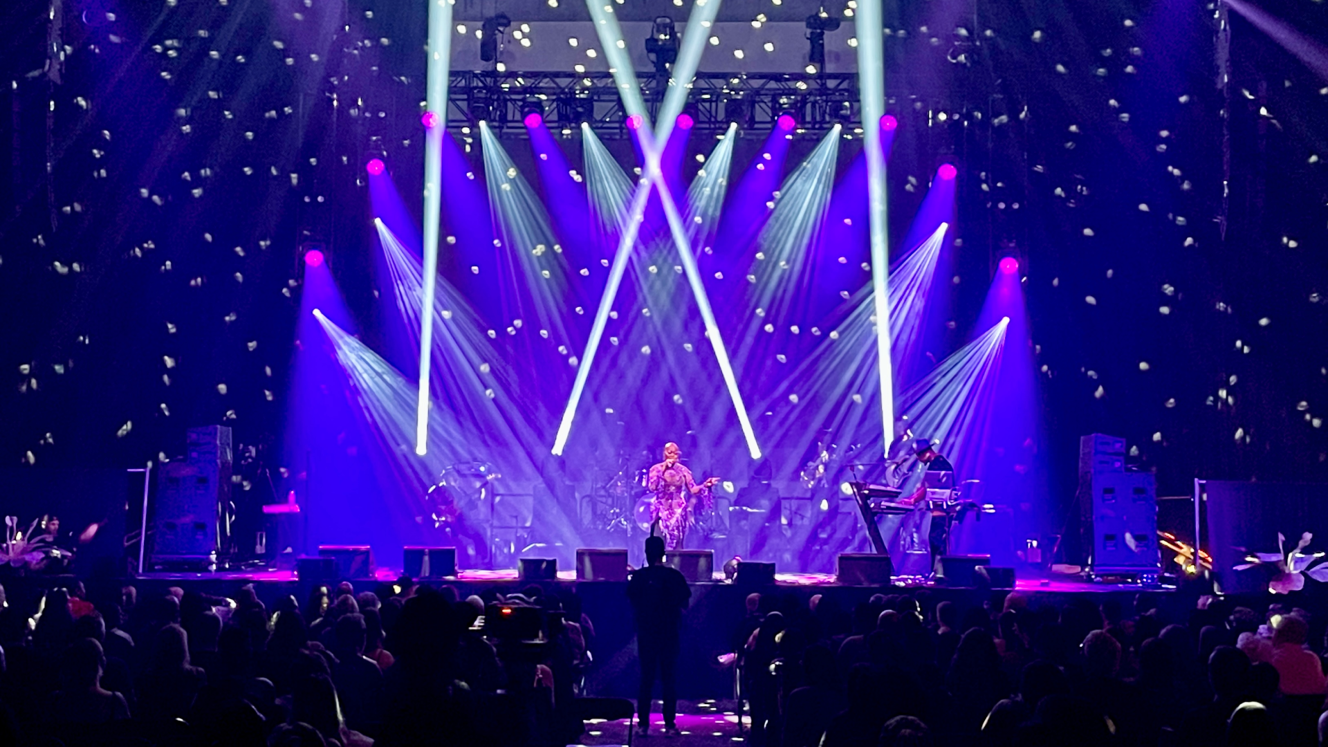Paisley Park Celebration Dark event stage illuminated by a low magenta LED strip at floor level, overhead moving-head lights casting amber and white beams upward, and intricate blue-and-gold gobo patterns projected across the stage floor in a high-impact lighting design