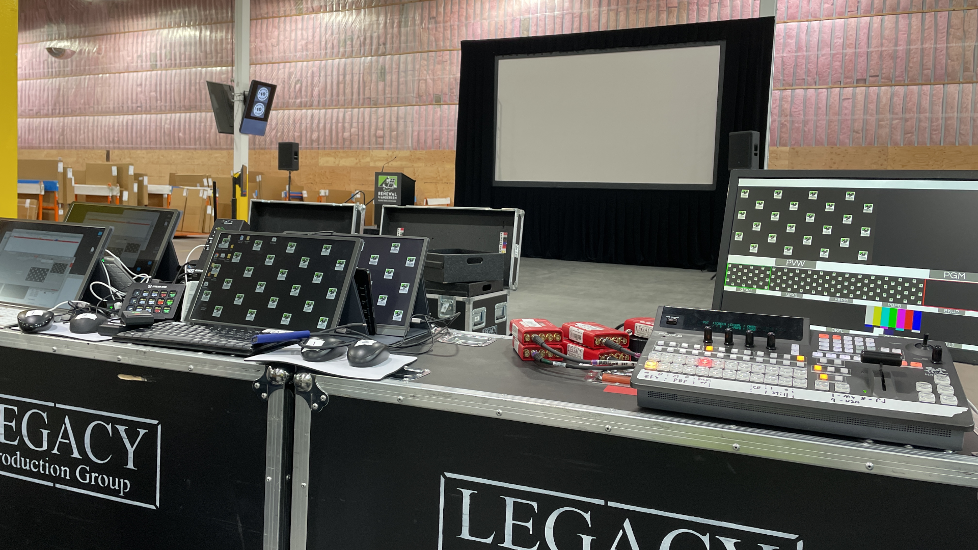 FOH Warehouse Event Live production control room with five operators wearing headsets seated at desks loaded with laptops, video switchers, and audio mixers; two large wall-mounted screens display multi-view camera feeds and a bright studio preview in a carpeted office space