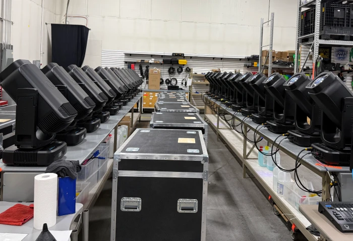 Warehouse staging area with two long workbenches each lined with black moving-head LED wash fixtures, central rows of black flight cases ready for transport, and industrial shelving of AV gear in the background, showcasing Legacy Production Group’s lighting inventory organization